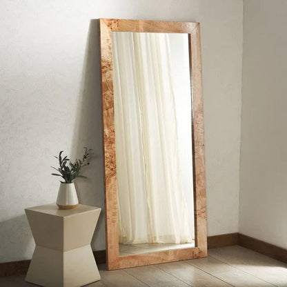 Wooden floor mirror with a white vase and plant on a small table against a plain wall.