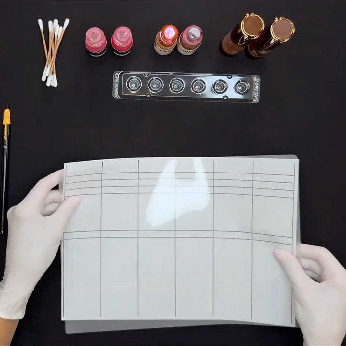 Gloved hands holding a pigment swatch grid with PMU pigments, pigment cup tray, and cotton swabs.