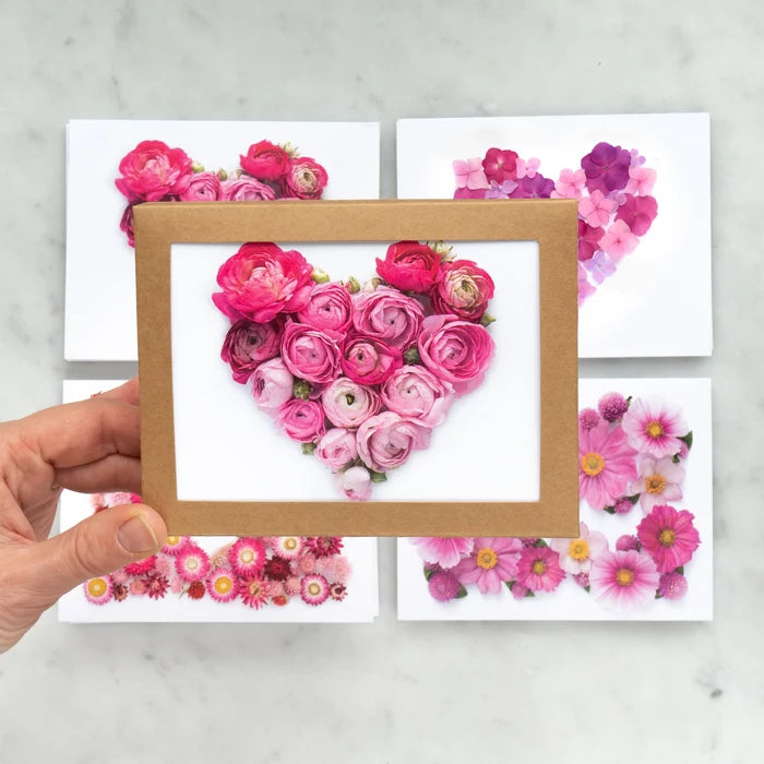 Heart-shaped arrangement of pink flowers in a wooden frame held by a hand on a light gray background.