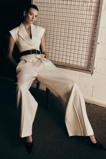 A model wearing natural beige mid-rise wide leg linen pants with a belt, sitting against a tiled wall.