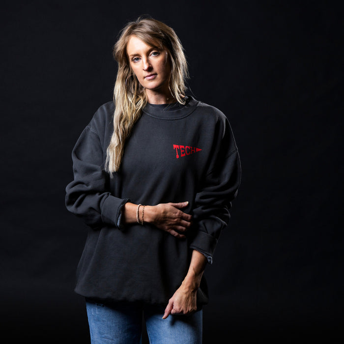 A woman modeling a black crewneck sweatshirt with a red 'TECH'  pennant logo on the chest, paired with blue jeans, set against a solid black studio background.